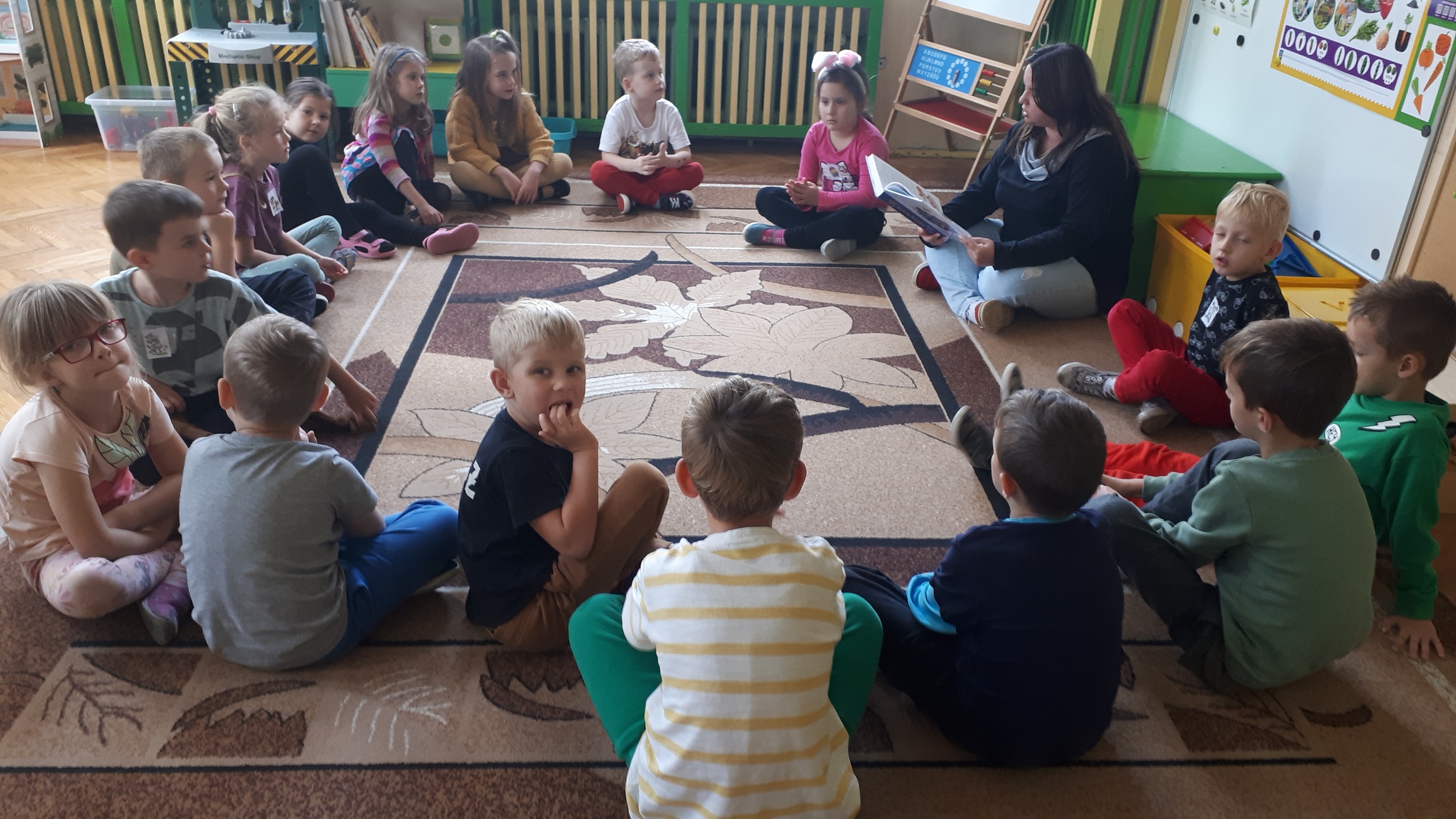 Na zdjęciu dzieci siedzą w kole na dywanie i słuchają nauczycielki czytającej książkę